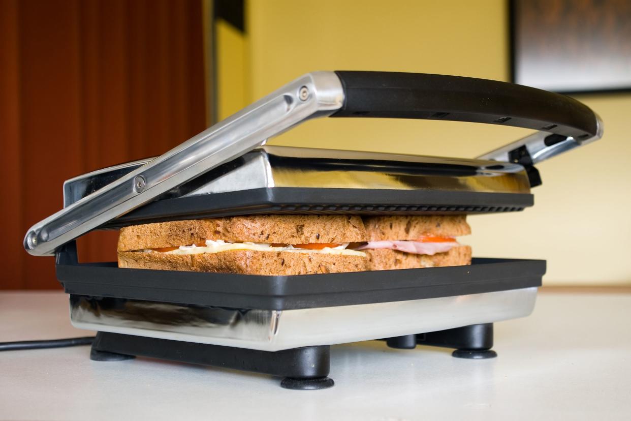 Closeup of an electric stainless steel sandwich grill with a sandwich inside, about to be pressed to cook it, on a countertop in a kitchen with a blurred background of a light yellow wall