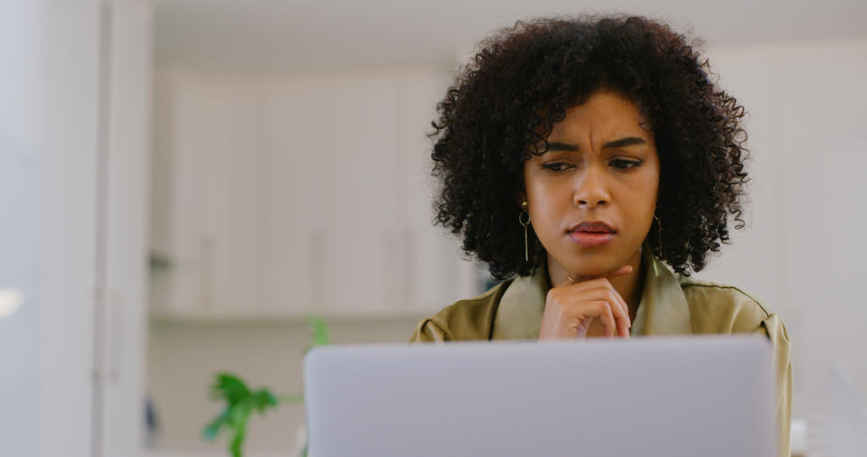 A woman with curly hair looks thoughtfully at a laptop in a home setting. She appears to be concentrating on something on the screen
