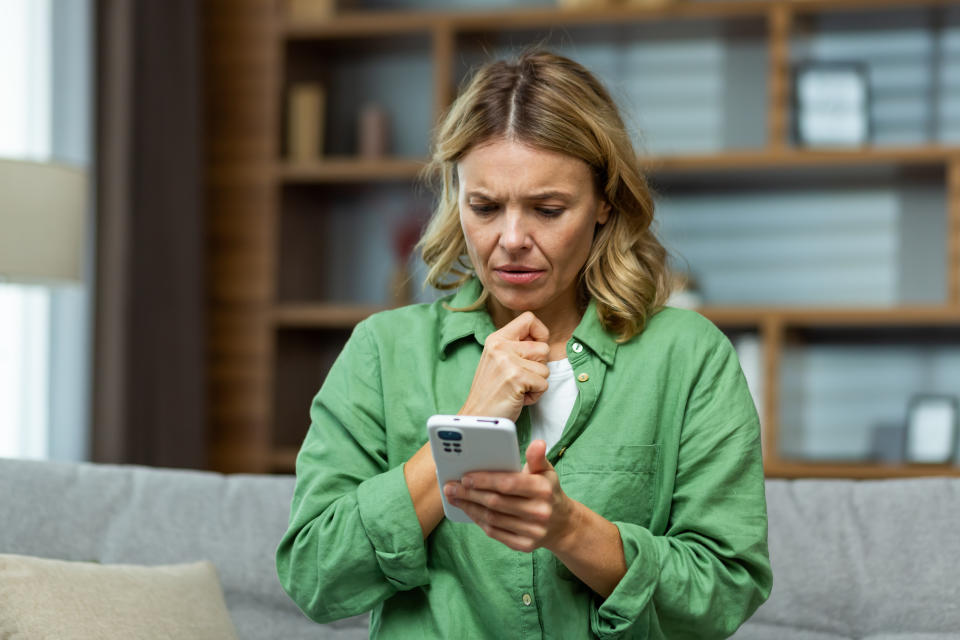 Upset senior woman sitting on sofa at home and holding phone. She looks worriedly at the screen, received a message, bad news.