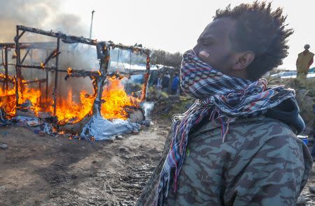 Migrants stand near a burning makeshift shelter set ablaze in protest against the partial dismantlement of the camp for migrants called the "Jungle", in Calais, France, March 3, 2016. REUTERS/Yves Herman