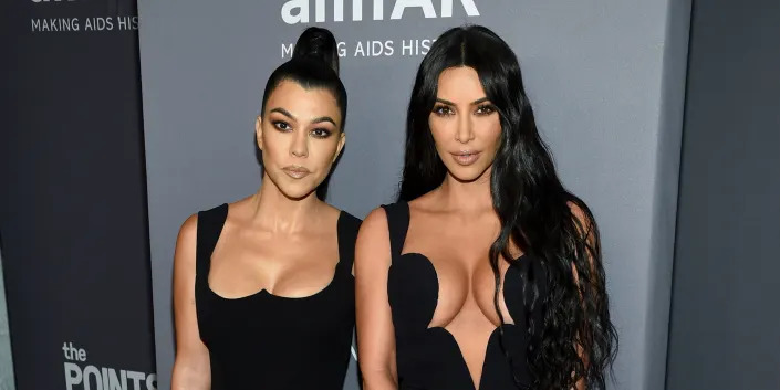 Kim and Kourtney Kardashian standing next to each other in front of gray background.