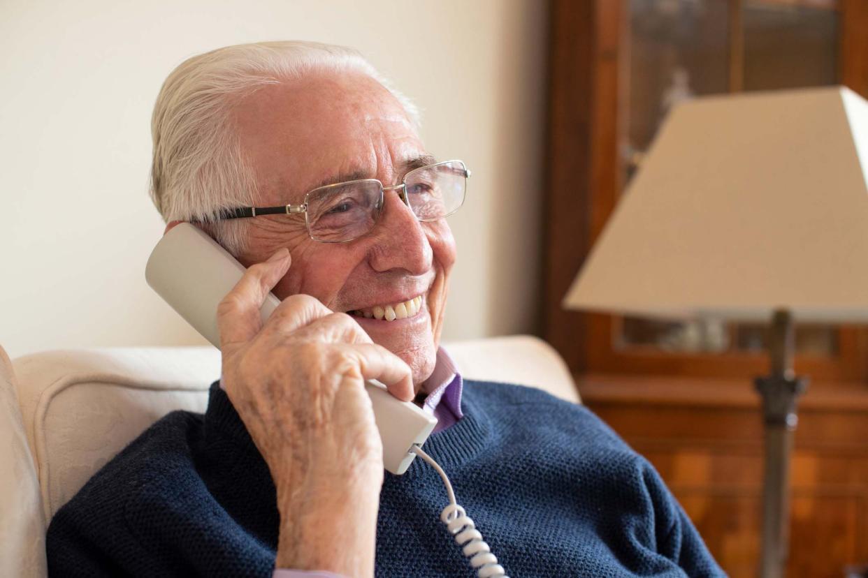 Side view of a senior man's face smiling while listening to a beige landline phone receiver, sitting on beige couch, with antique wood furniture against the wall and standing lamp in the background