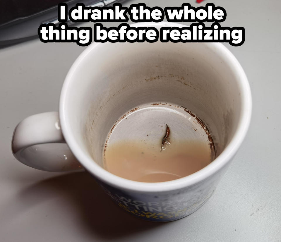 A partially full coffee mug with a used teabag inside it, placed on a desk