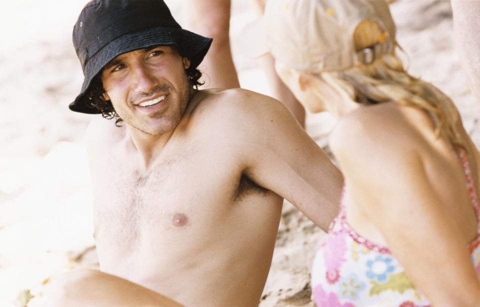 A shirtless man wearing a bucket hat and a woman in floral swimwear are sitting on the beach, smiling at each other