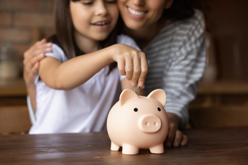 Close up smiling mother and little daughter hugging, putting coin into pink piggy bank, caring mum teaching adorable girl child to saving money for future, insurance and investment concept