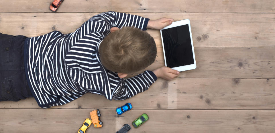 A boy lying on the floor using a tablet computer with toy cars scattered around him.