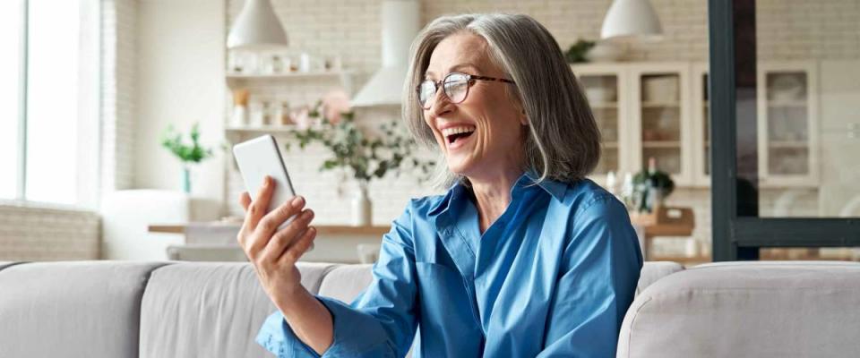 Happy mature old 60s woman sitting on couch and holding smartphone