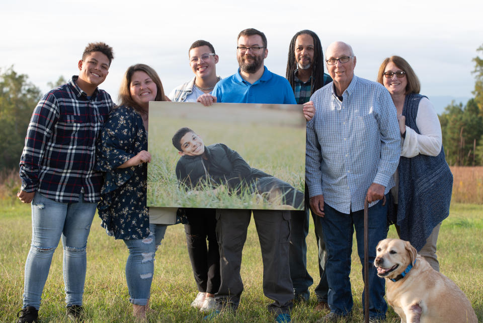 The family and John Wilson, holding a large poster of Keegan, line up in a meadow for a photo shoot with a family dog.