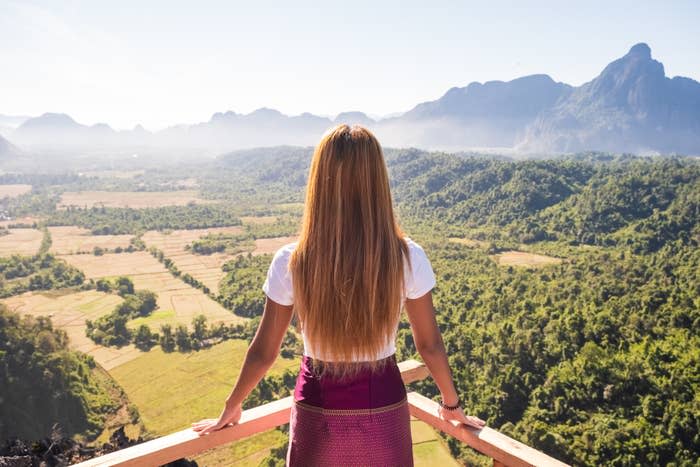 A woman with long hair stands on a wooden balcony overlooking a vast, green valley with mountains in the background