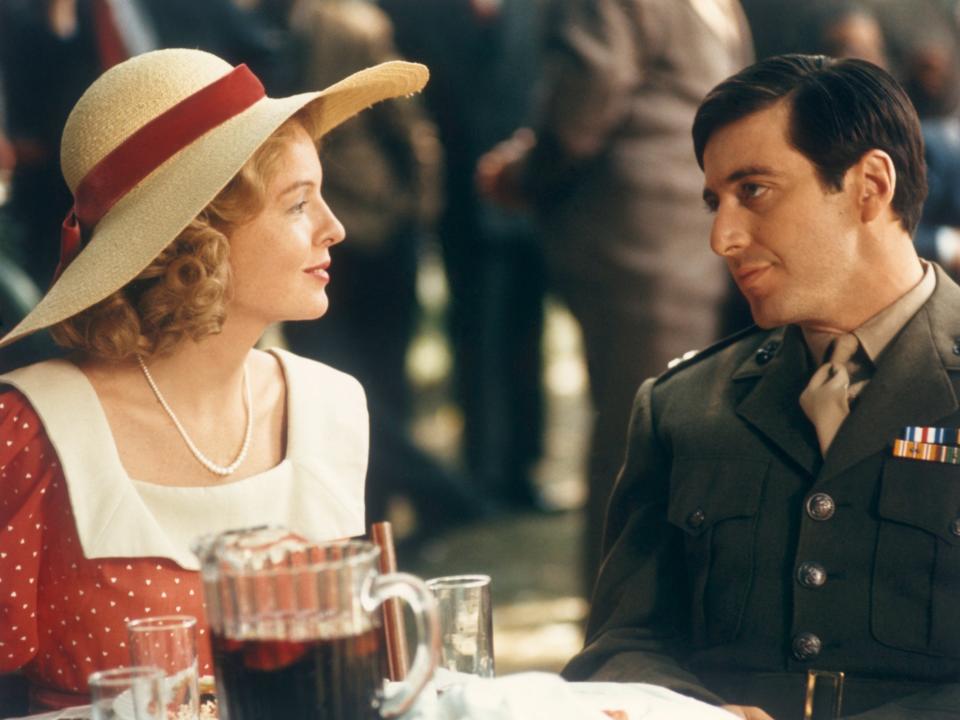 Keaton and Pacino in character sitting at a table. Kay is in a red and white dress and sunhat and Michael in a military uniform.