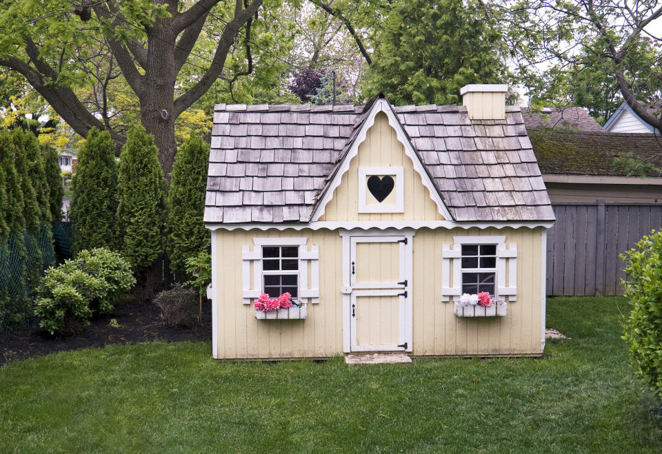 A wooden pale yellow play house with flowers on the windows