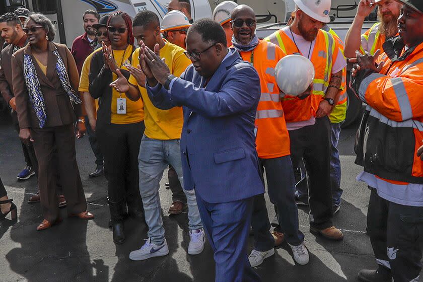 A man claps, with people, some in orange vests, behind him.