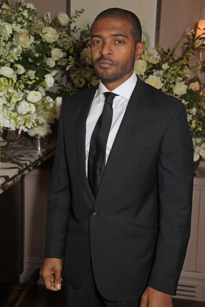 Man in a black suit and tie standing indoors with floral arrangements in the background. Name not provided