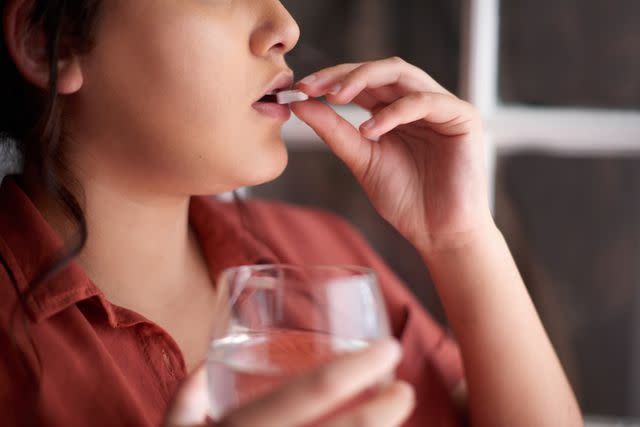 <p>Tassii / Getty Images</p> An individual holds a glass of water and takes a supplement.