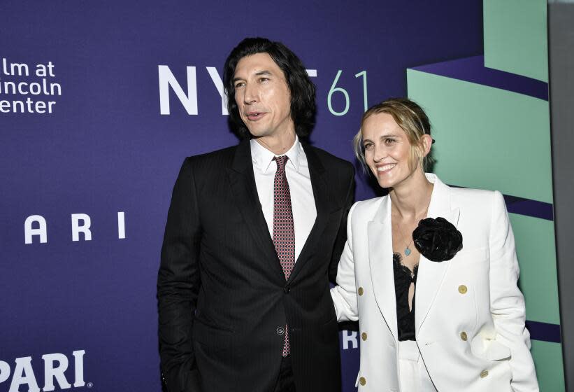 Adam Driver in a black suit stands next to Joanne Tucker in a white suit as the pair smile and embrace