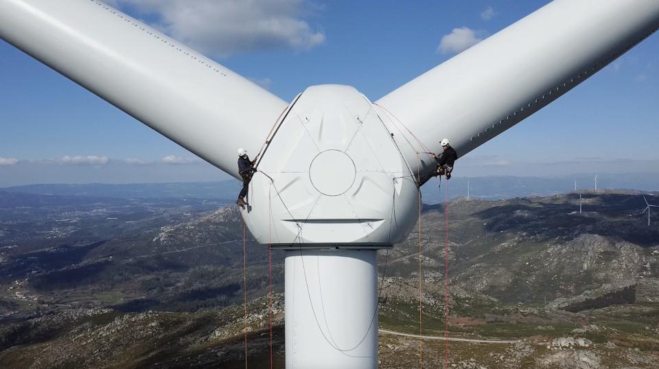 Two wind turbine technicians hang from cables on the side of a wind turbine.