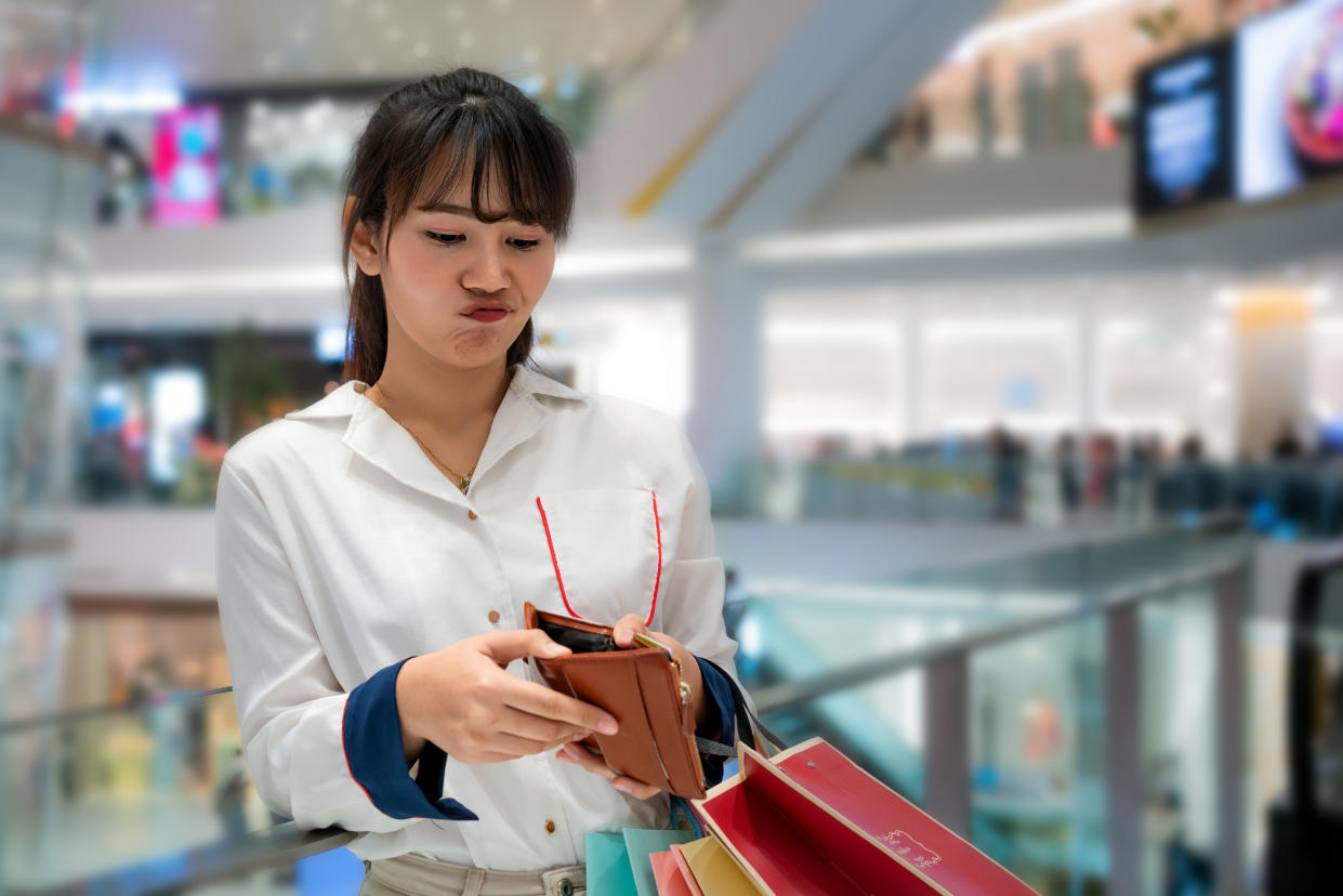 An Asian woman stands in a shopping mall with a frustrated look on her face as she looks inside her empty purse, while carrying several shopping bags