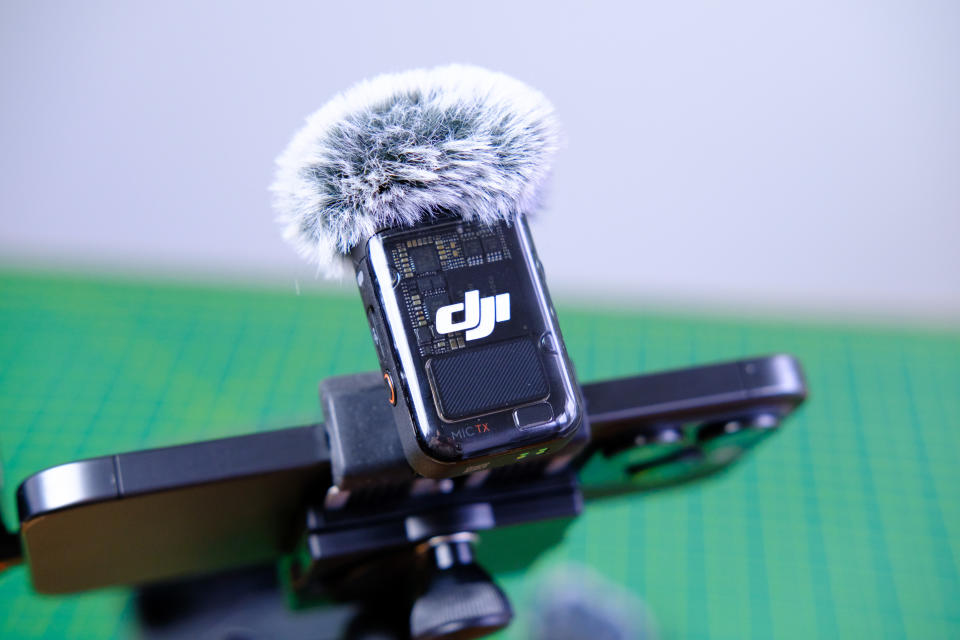 A photo of the DJI Mic 2 (TX unit) in use with an iPhone 15 Pro. The TX unit is mounted on top of the iPhone which is horizontally aligned for filming.