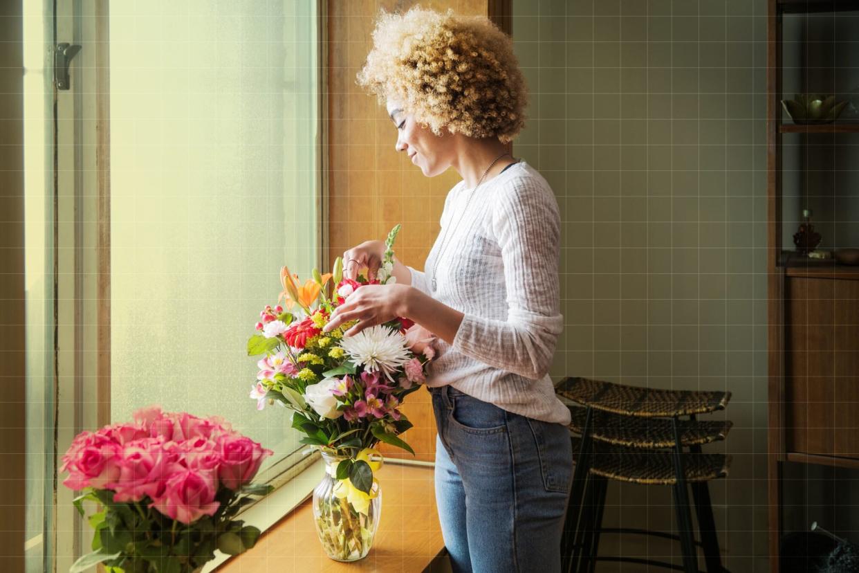 Smiling young woman arranging flowers in container on window sill at home