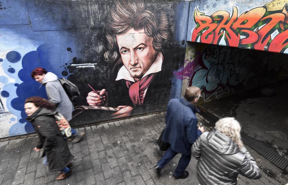 People passing a graffiti showing composer Ludwig van Beethoven on a street in Bonn, Germany, Wednesday, Feb. 19, 2020. The city of Bonn, where Beethoven was born on December 17, 1770, celebrates the 250th birthday of the great German composer with many cultural events during this year. (AP Photo/Martin Meissner)