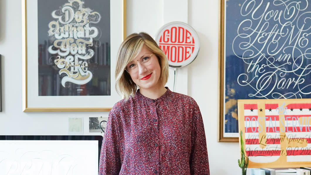  Jessica Hische headshot, woman in a red top in front of her lettering work. 