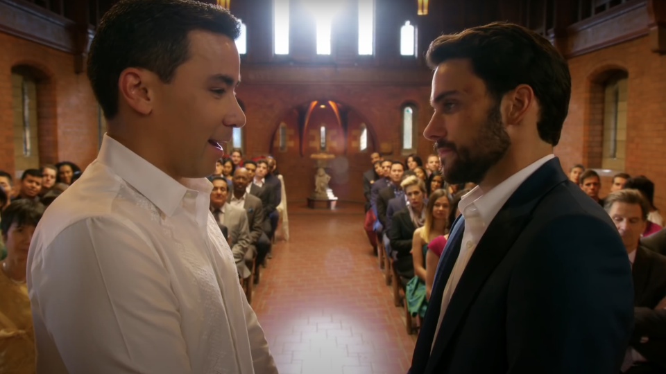 Two men, one in a white shirt and the other in a navy suit, are standing at the altar of a church, preparing to get married, with guests seated behind them