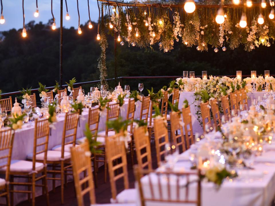 Long tables set up for a wedding. Lights hang overhead.