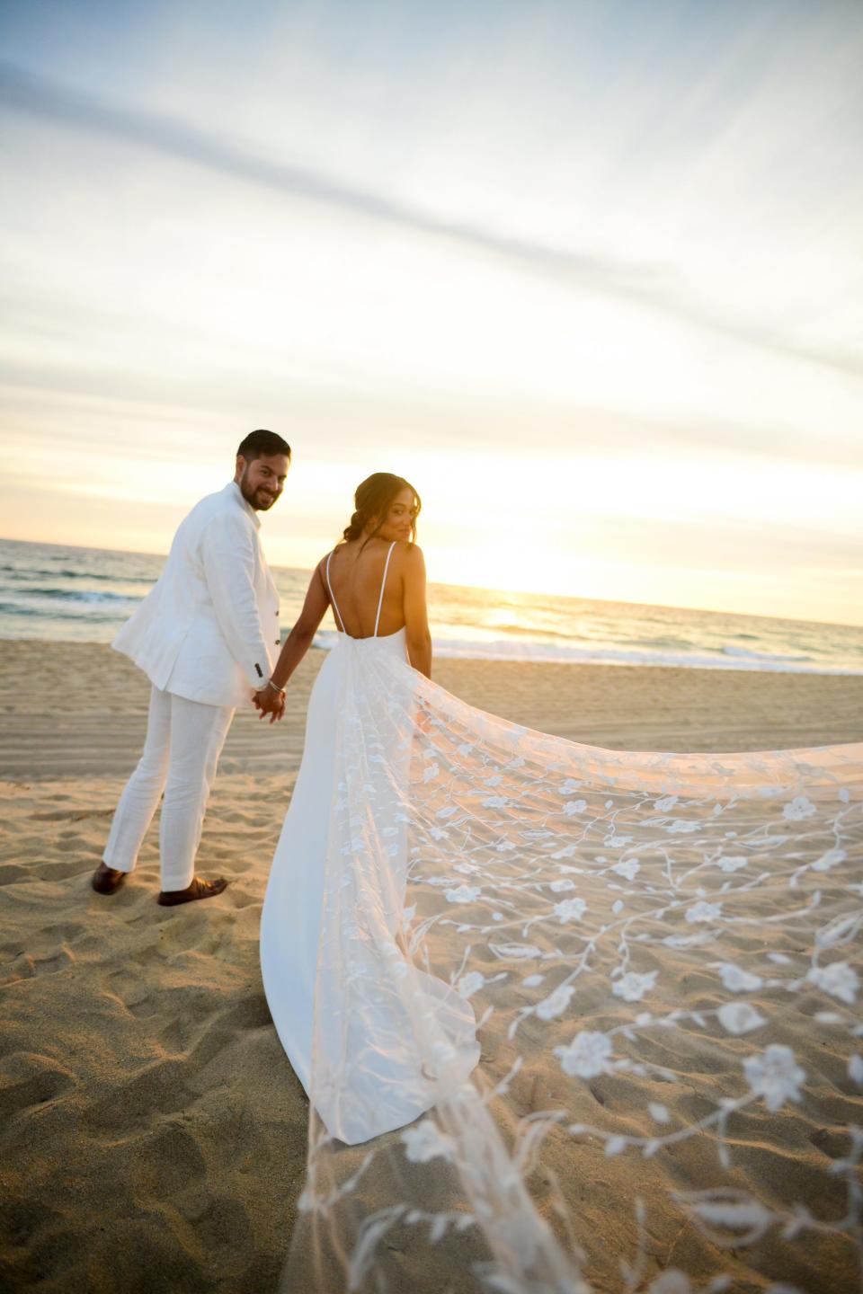 A man and woman look over their shoulders in their wedding attire on a beach.