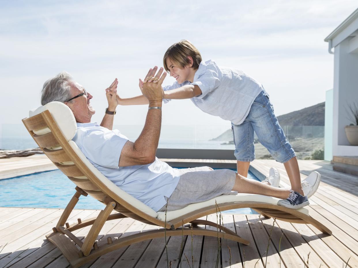 An old man lying on a recliner by a pool plays with his grandson