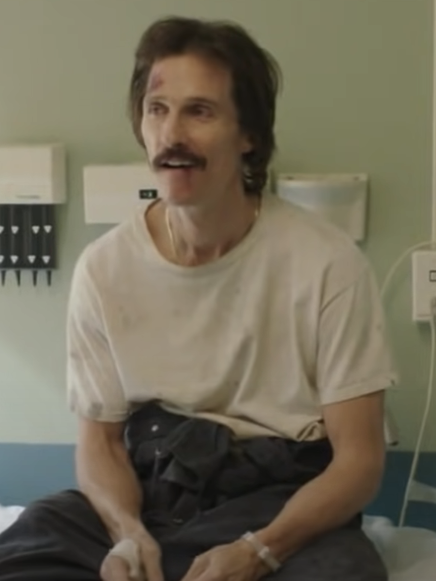 Matthew McConaughey sits on a hospital bed in a plain t-shirt and pants, with visible facial bruises and a mustache