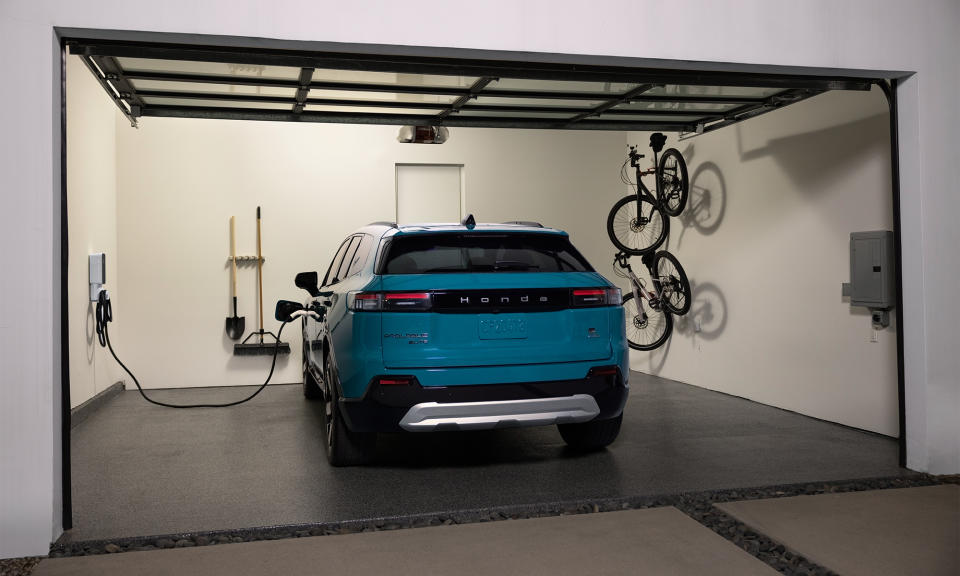 Honda marketing photo of the Prologue electric SUV charging in a home garage. Two bicycles hang on wall-mounted racks to the vehicle's right.