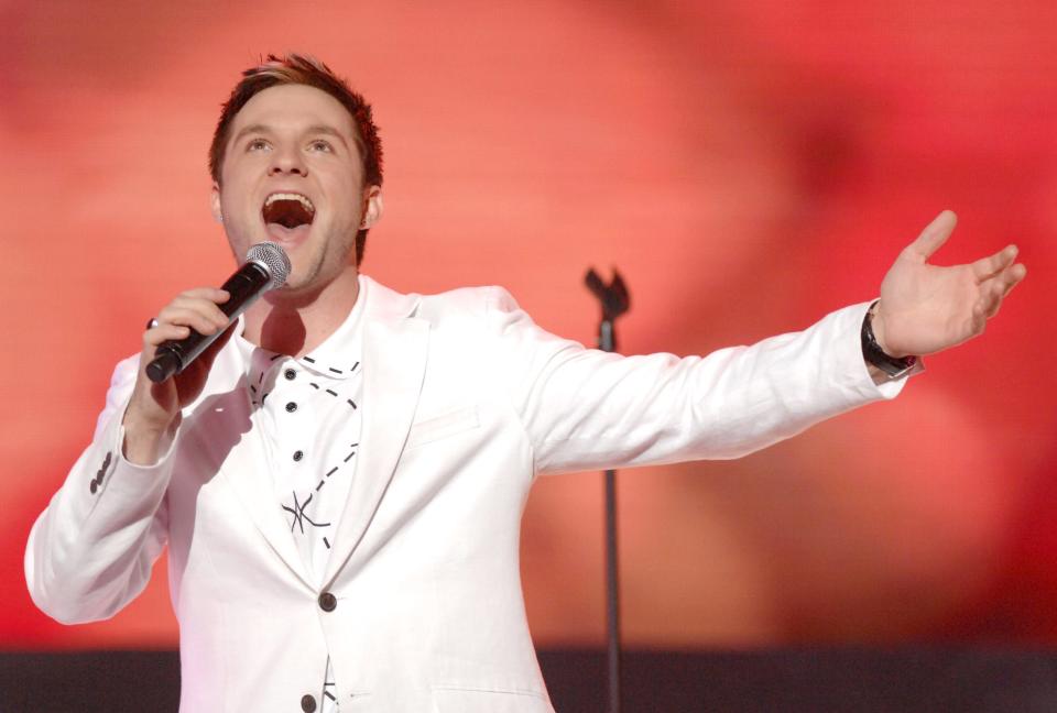 Blake Lewis wears a white suit and holds a microphone and sings in front of a red background