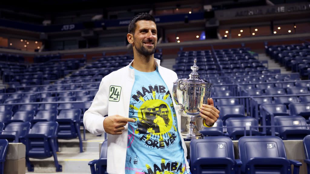 novak djokovic poses for a photo in empty stadium stands, he smiles and looks off camera while holding a silver trophy in one hand and pointing to his blue t shirt with the other hand, he also wears a white jacket with the number 24 on the front and blue shorts