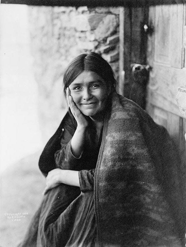 Photo shows a Navaho woman sitting outside doorway, right hand on cheek, facing front, smiling.