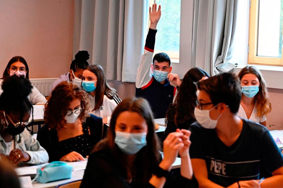 A masked high-school student in the back of a crowded classroom, where everyone is wearing masks, raises his hand.