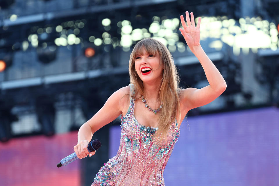 Taylor Swift performing on stage in a sparkly dress with a microphone in hand, waving to the audience