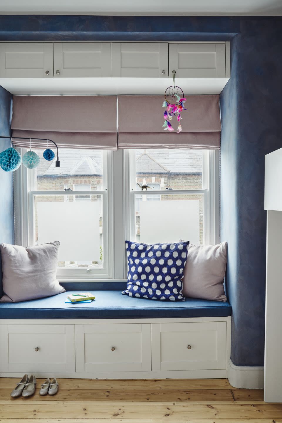 a windowseat in a childs bedroom with built in storage the walls are painted deep blue and the windowseat is accessorised with pink and blue cushions and a pink blind