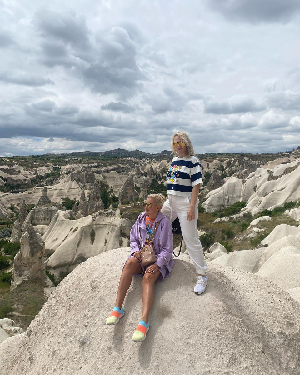 Rebel Wilson wears a black and white striped top with white pants, standing on a rock next to her girlfriend Ramona Agruma in a lilac outfit sitting on the rock.