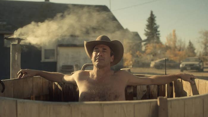 Man relaxing in a rustic wooden hot tub, wearing a cowboy hat, with steam rising in a rural setting