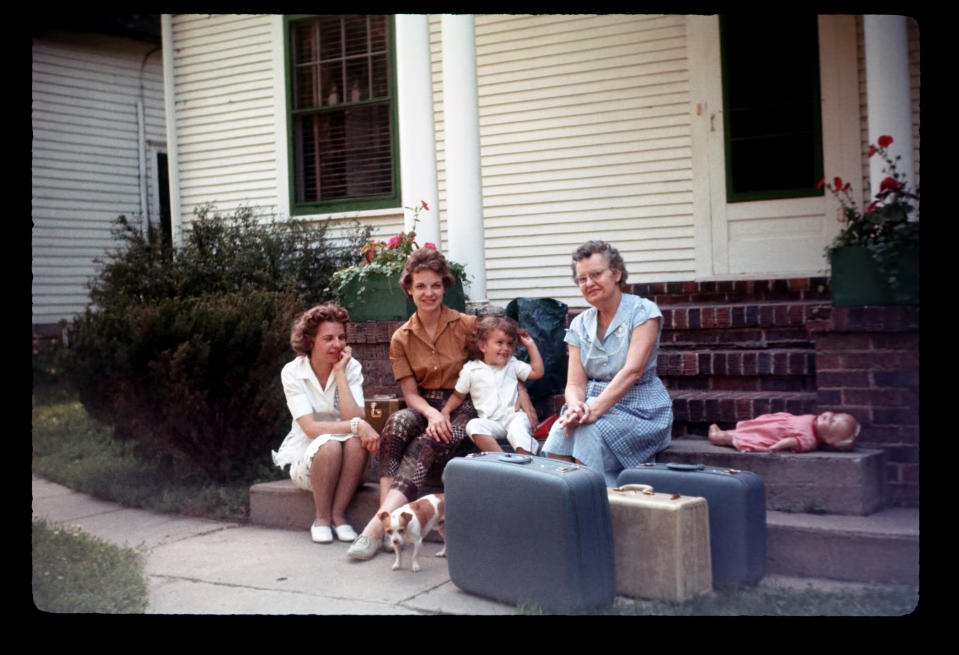 Three women and a little girl sit on steps outside a house, with suitcases, a doll, and a small dog. The women wear casual 1950s attire