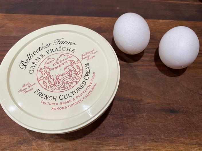 Can of cr&#xe8;me fra&#xee;che next to two eggs with white eggshells on wooden cutting board