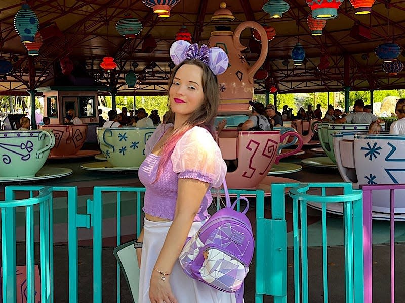 The writer stands in front of a ride at Disney World with tea cups. The writer wears a white skirt, purple shirt, mouse ears, and a purple backpack