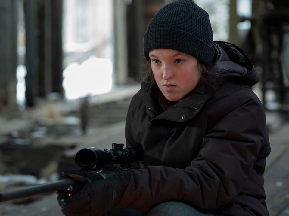 ellie, played by bella ramsey, in the last of us. she's wearing a winter coat and black beanie, crouching in a decrepit building and holding a rifle. there's an intent expression on her face
