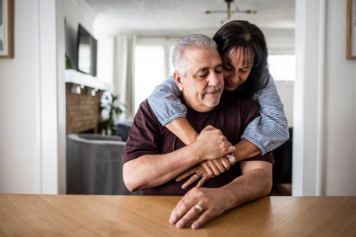 A couple shares an intimate moment at home; the woman lovingly hugs the man from behind as they sit at a wooden table