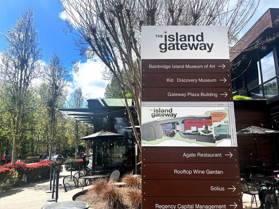 A white and brown sign with a map of the island gateway on Bainbridge Island in front of a small shop and seating area with trees and a blue sky in the background