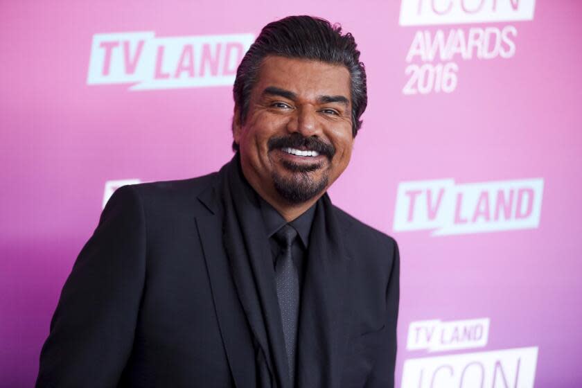 A man with a mustache, a beard and dark hair smiling and posing in a black suit and tie