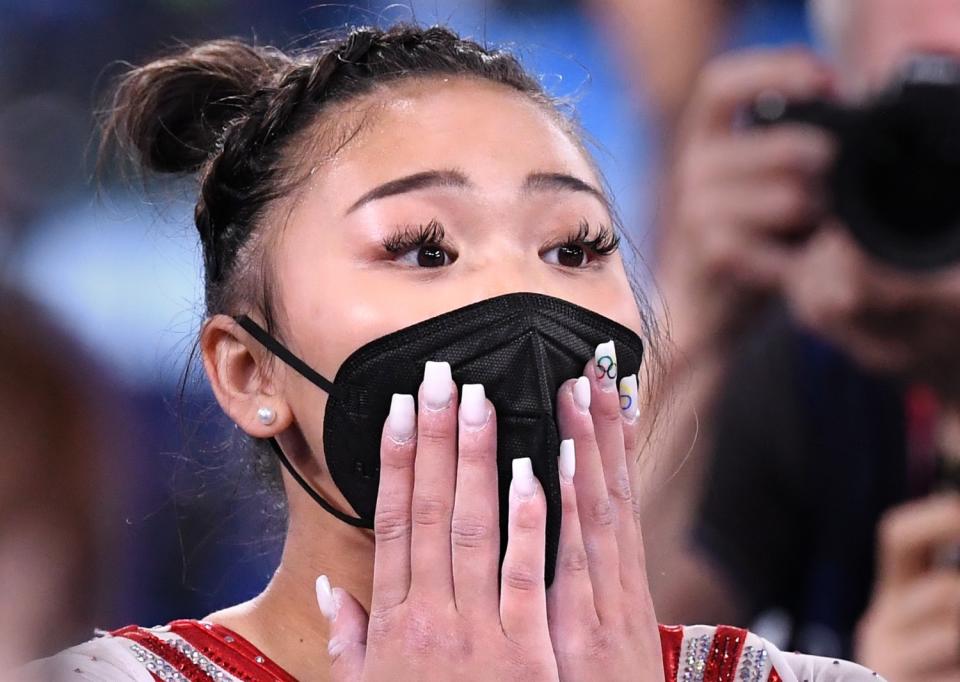 Suni Lee, wearing a mask, holds her hands to her face