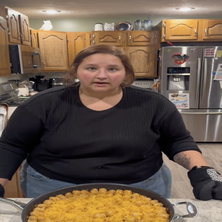 a tiktok creator standing in front of a large skillet that is lined with tater tots