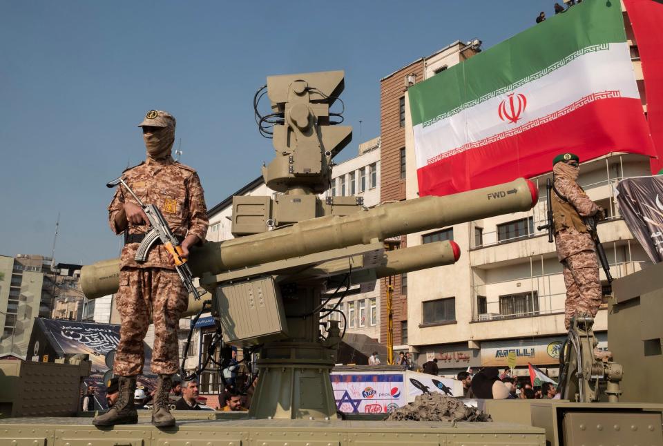 Military guards stand next to an antiaircraft missile system with the Iran flag on a building in the background.