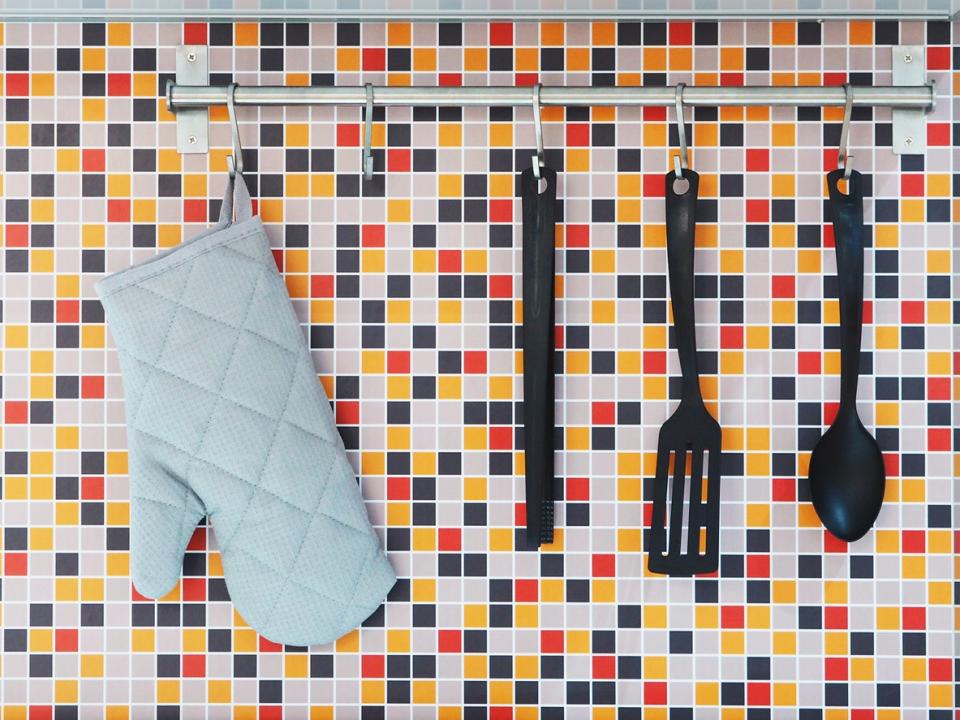 Black kitchen utensils hanging next to blue oven mitten in front of colorfully tiled kitchen wall